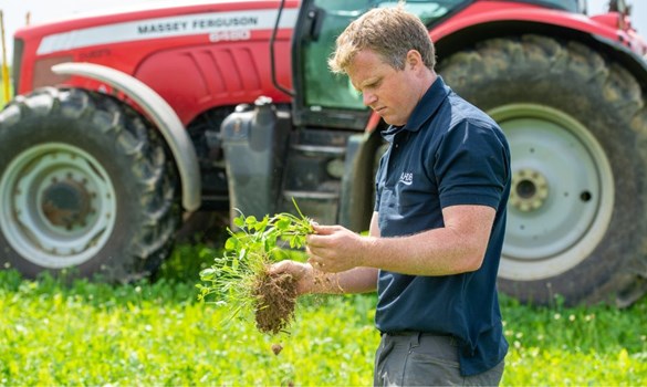 Man stood in grass in front of red tractor holding a handful of grass, soil and herbs